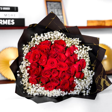 Bouquet of red roses with white baby’s breath wrapped in black and gold paper on display indoors