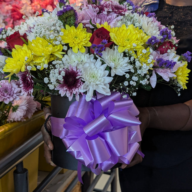 Bloomy hatbox with mixed flowers including chrysanthemums, roses, and fillers arranged in a black box with purple ribbon.